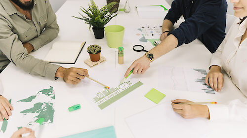 Vier Personen sitzen um einen Tisch herum, auf dem verschiedene Papiere mit Grafiken zum Thema Nachhaltigkeit liegen. Zwei der Personen deuten auf ein in der Tischmitte liegendes Papier mit einer Abbildung zum Energiesparen.