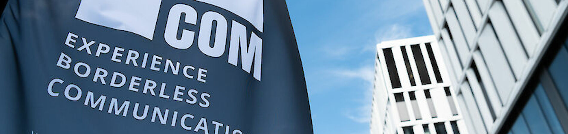 Beachflag mit Logo und Schriftzug BOCOM vor blauem Himmel und Gebäuden im Hintergrund