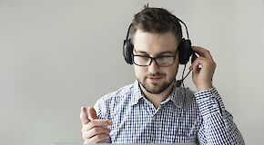 Young man with beard and glasses wears headset and looks at laptop screen