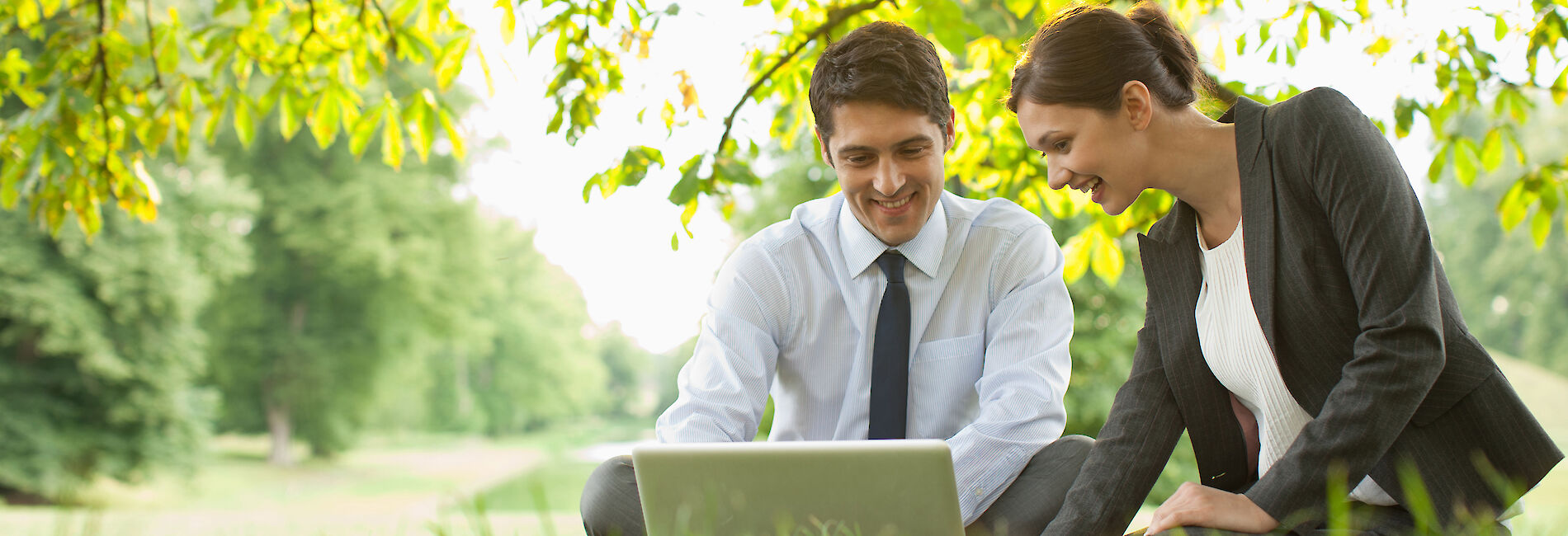 Man and woman in business outfits sitting on a meadow with a laptop in front of them.