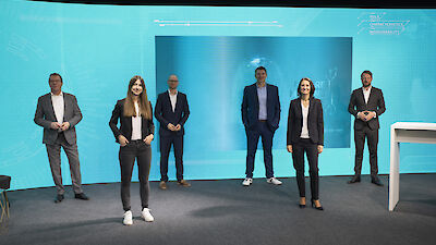 Sechs Personen stehen mit jeweils ca. anderthalb Metern Abstand voneinander in einem Studio vor einem petrolfarbenen Hintergrund mit Grafik