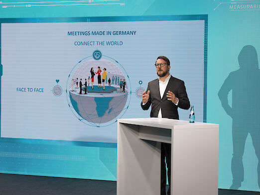 Matthias Schultze giving a presentation, with a graphic on a large screen in the background.