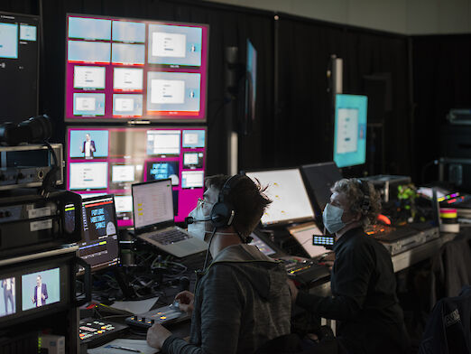 Backstage view into the technical area of the streaming studio with numerous monitors and computers as well as two employees with mouth-to-nose coverings.