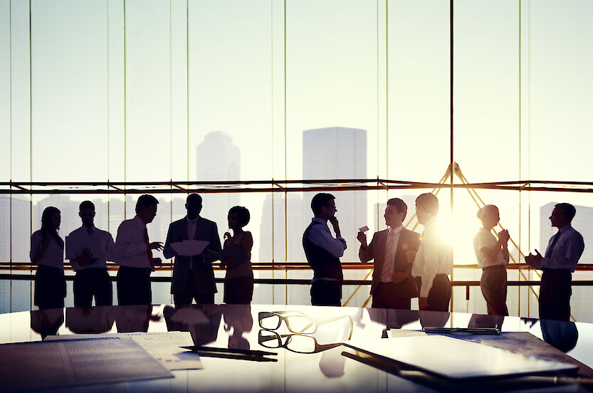 Ten people in business outfits stand in a meeting room in front of a glass facade