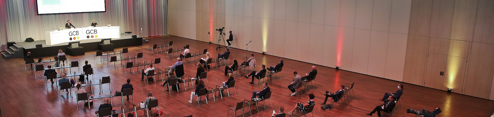 Ansicht eines Saals von oben, in dem ca, 30 Menschen mit großen Abständen auf Stühlen sitzen und nach vorne in Richtung einer Bühne mit Leinwand blicken.