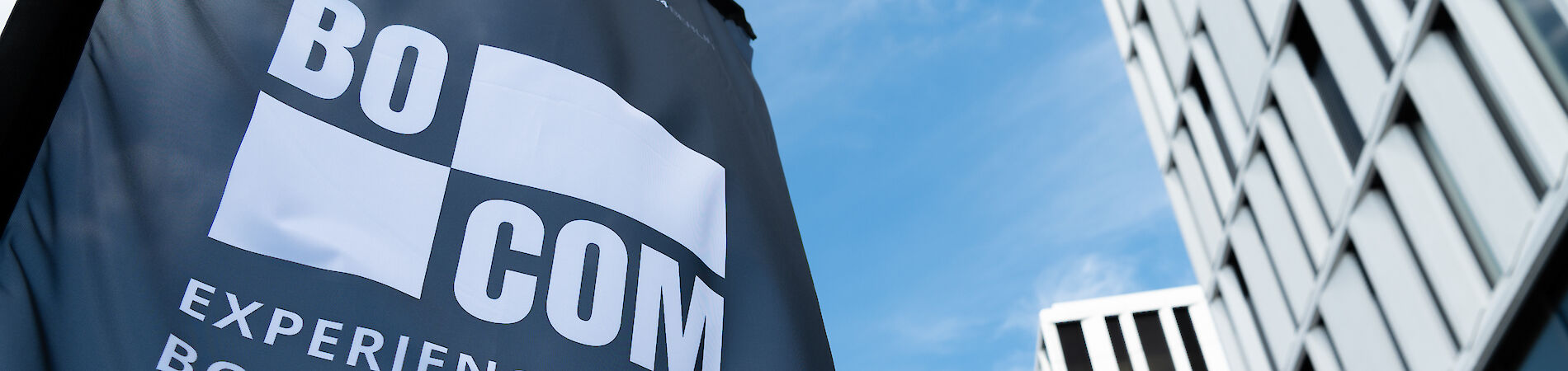 Beachflag mit Aufschrift BOCOM - Experience Borderless Communication, im Hintergrund ein Gebäude und blauer Himmel