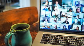 Auf einem Tisch steht ein Laptop. Auf dem Bildschirm sind verschiedene Teilnehmer eines Online-Meetings zu sehen.