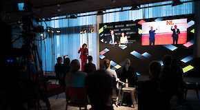 Teilnehmer*innen einer Konferenz im Publikum, Referenten digital auf der Leinwand zugeschaltet.