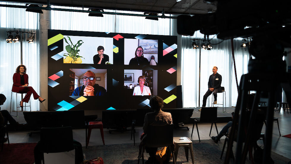 Im Konferenzraum wird ein virtueller Vortrag von vier Referenten, die jeweils auf einem Bildschirm eingeblendet werden, verfolgt.