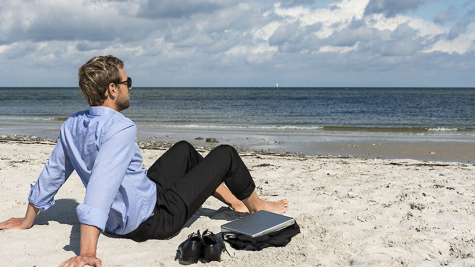 Mann in Business-Outfit sitzt Barfuß am Strand, neben ihm ein Laptop und seine Schuhe.