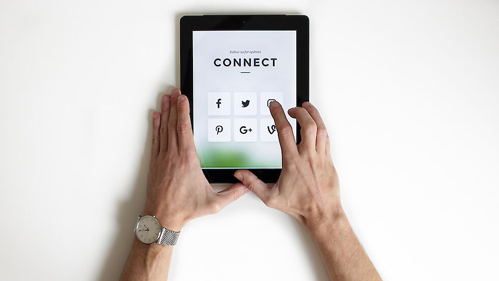 Zwei Hände halten ein Tablet. Ein Finger berührt den Bildschirm, auf dem mehrere Social Media-Icons zu sehen sind. | © Nordwood Themes / Unsplash.com