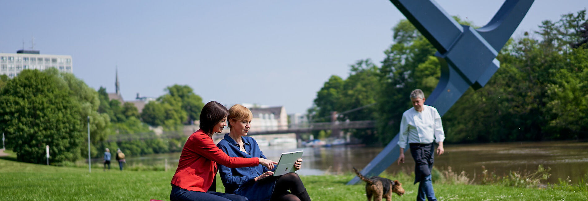 Zwei Frauen sitzen auf roten Würfeln im Gras an einem Flussufer, im Hintergrund ist die Spitzhacke Kassel zu sehen.