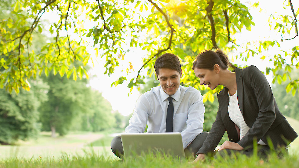 A man and a woman in business outfits sitting on a meadow with a laptop in front of them.